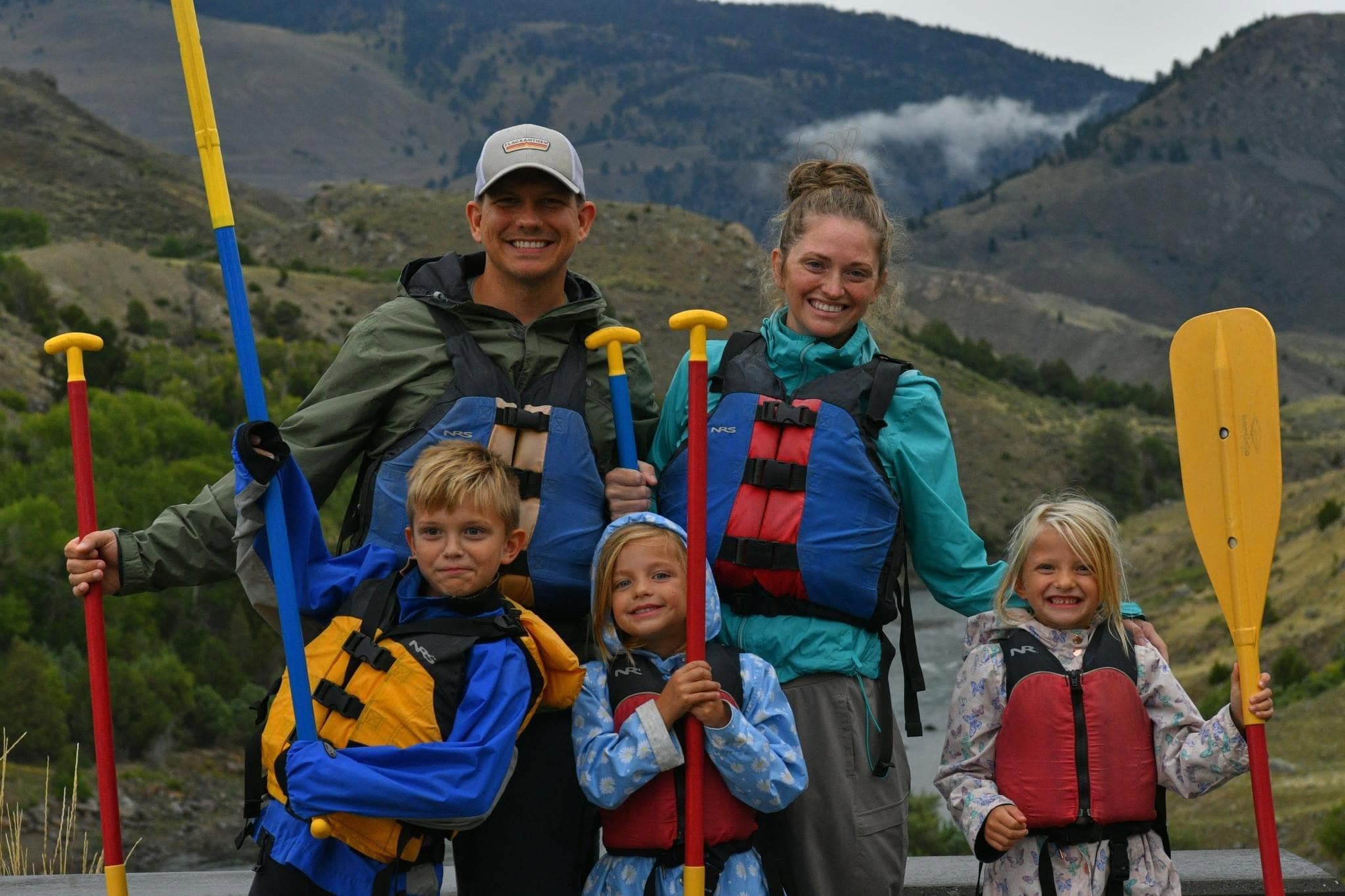 Josh and Johanna Ackerman with Harrison, Everly, and Emma in life jackets holding rafting paddles by a river in the mountains